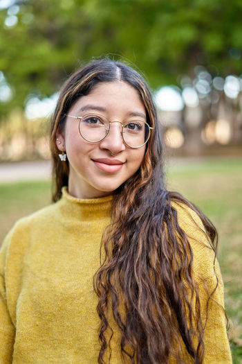Content young female in eyeglasses looking at camera while sitting on grassy lawn in park with trees on blurred background