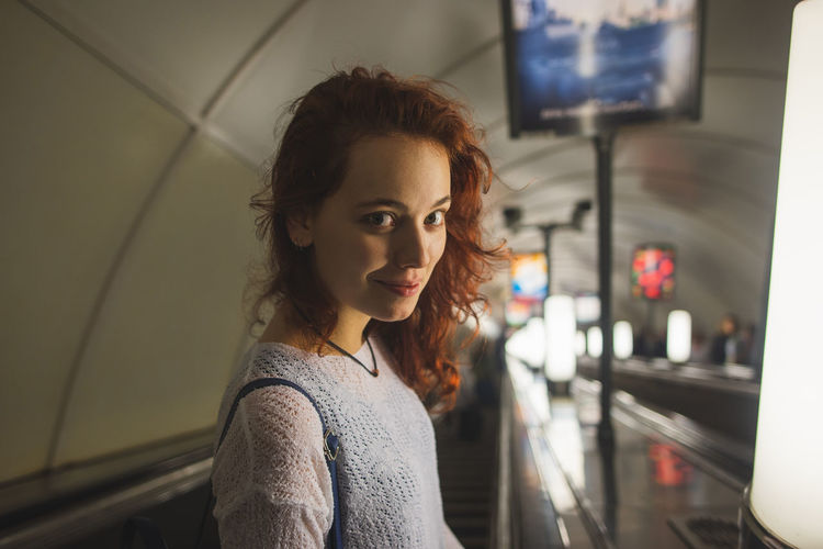 High angle side view of young female passenger in casual outfit with red curly hair looking at camera with smile while going down subway escalator in saint petersburg