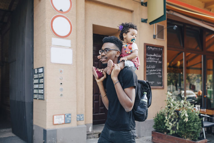Young man carrying daughter on shoulders while walking by building