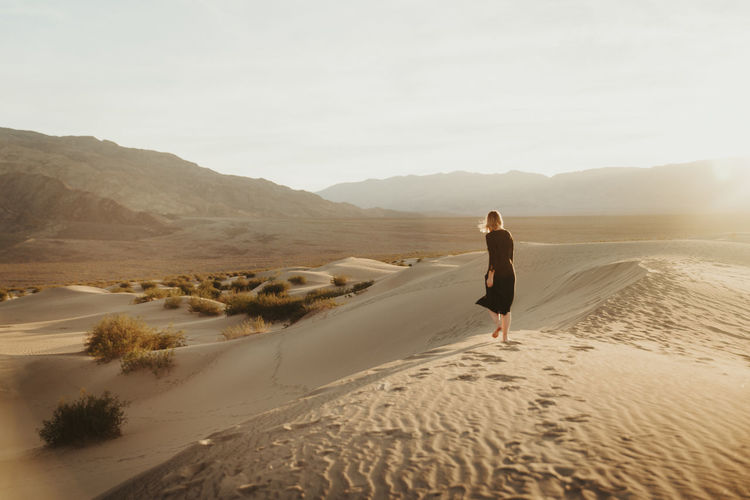 Rear view of woman walking at desert against sky during sunset