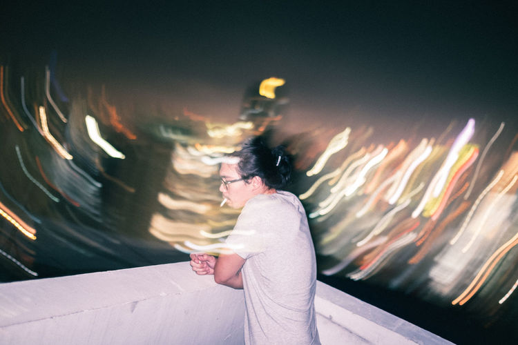 Side view of man smoking while standing on terrace against illuminated city at night