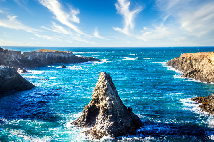 Cone shaped rock in the pacific ocean under a blue sky with clouds, california