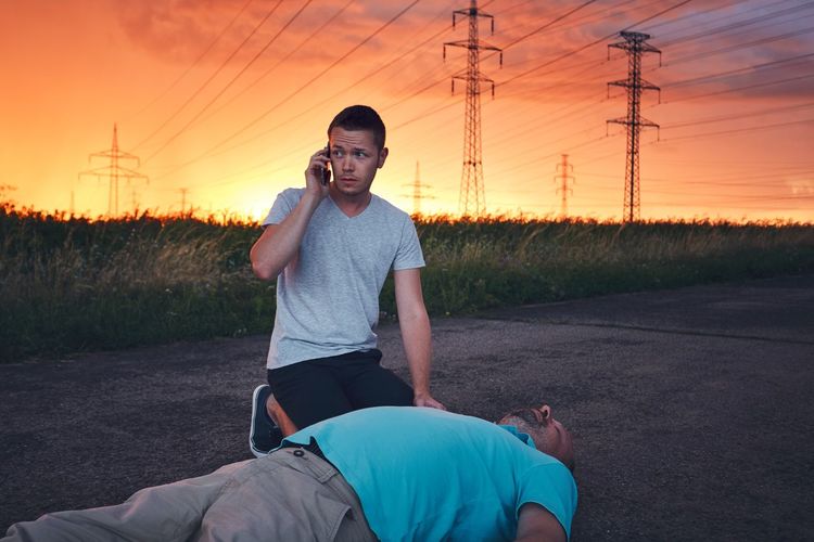 Man using mobile phone while unconscious friend lying on road against cloudy sky during sunset