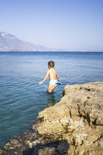 Full length of shirtless man standing on rock in sea against sky