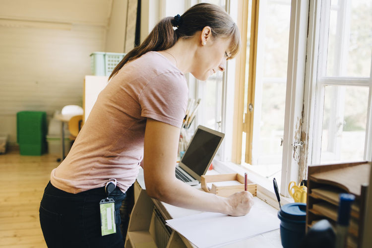 Confident teacher writing on paper while standing by window at classroom in school