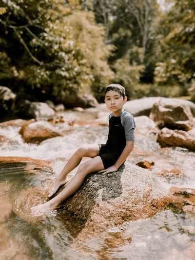 Full length portrait of boy sitting on rock in stream