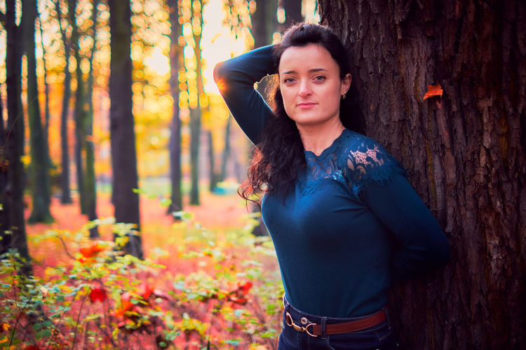 Portrait of beautiful young woman standing by tree trunk in forest during autumn