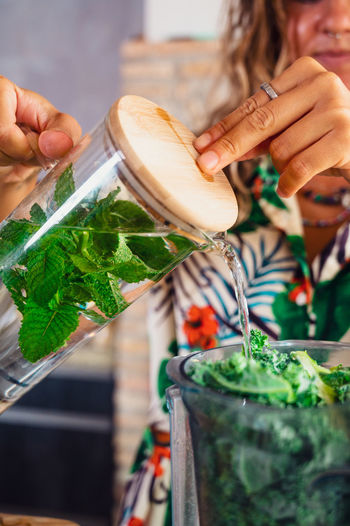 Crop unrecognizable female pouring refreshing water with mint into blender bowl with green kale while preparing smoothie in light kitchen