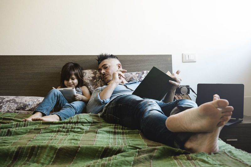 Full length of father and daughter holding book lying on bed