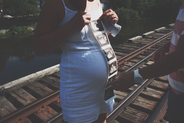 Midsection of pregnant woman with man holding instant print transfer while standing on railway track