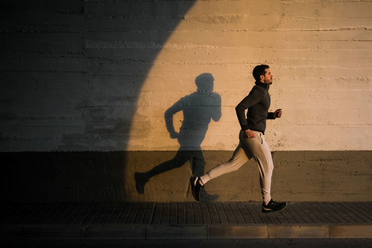 Side view of active guy in sportswear training legs jogging in urban street near a concrete wall