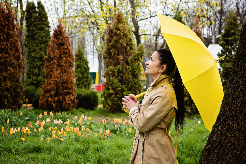 Happy senior woman in yellow rain coat with yellow umbrella walking in park. cheerful mature