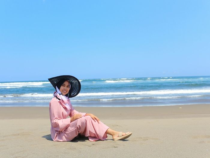 Woman wearing hat while sitting on beach by sea against sky