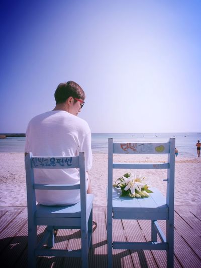 Rear view of man looking at lily flowers on wooden chair at beach against clear sky