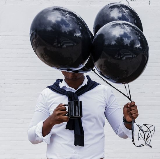 Man holding black helium balloons and coffee cup against white wall