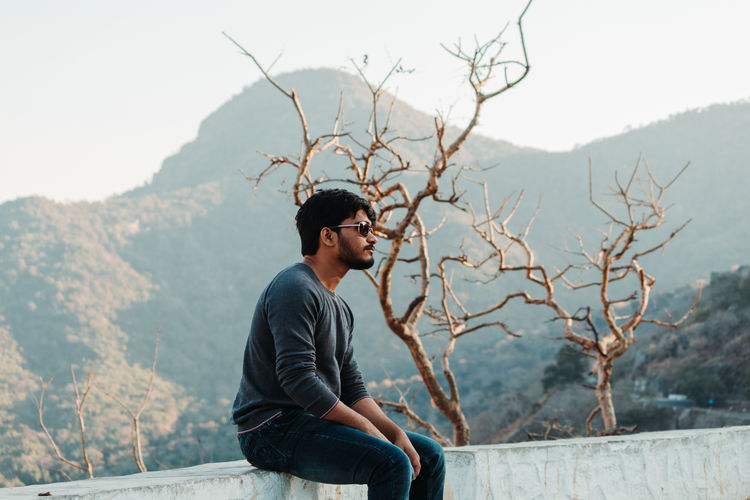 Young man sitting on retaining wall against trees and mountain