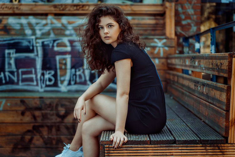 Portrait of beautiful young woman sitting on bench against graffiti wall
