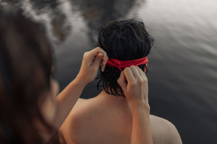 Crop anonymous female tourist tying red blindfold on head of partner against rippled water during trip
