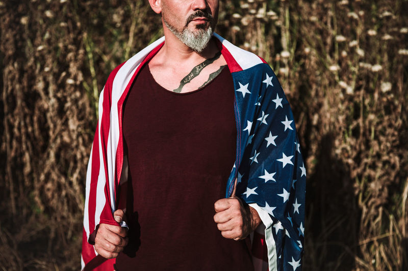 Serious cropped unrecognizable middle aged bearded male with tattoo wrapped in american flag looking away while standing against blurred nature