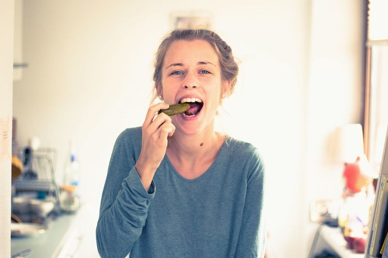 Portrait of happy young woman eating pickled cucumber at home