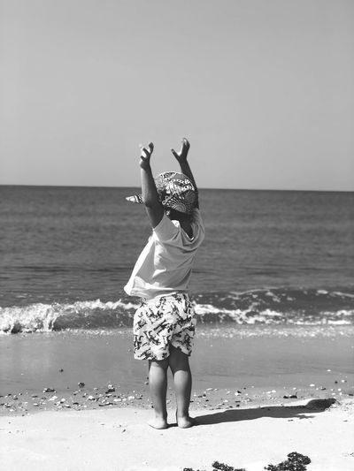 Rear view of boy with arms raised standing on beach against clear sky during sunny day