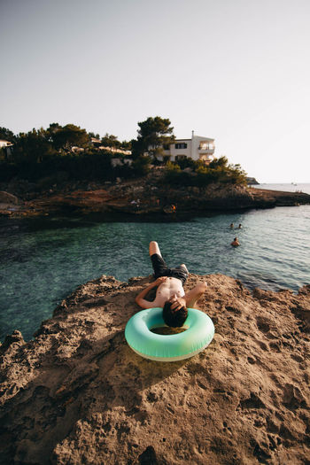 High angle view of shirtless mature man with inflatable ring lying on rock at beach against clear sky during sunset