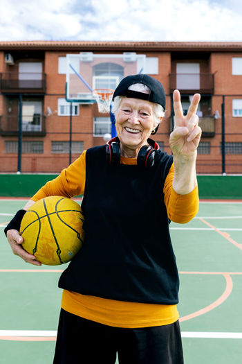 Optimistic mature female in activewear and headphones looking at camera while standing on public basketball court with ball during training