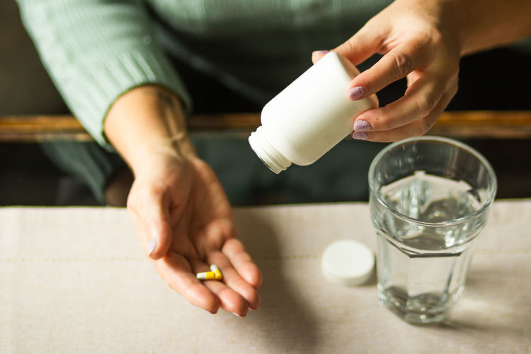 Elderly woman holding white plastic bottle and two capsules in hand, ready to take supplement pills.