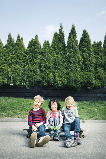 Portrait of happy children sitting on skateboard against trees
