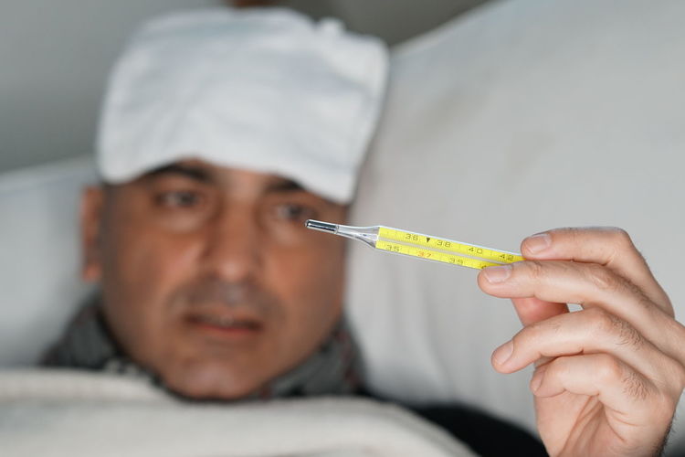 Close-up of man holding thermometer while relaxing on bed at home