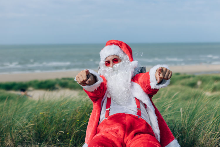 Santa claus sitting on the deck chair by the sea invites you to go on vacation pointing at you
