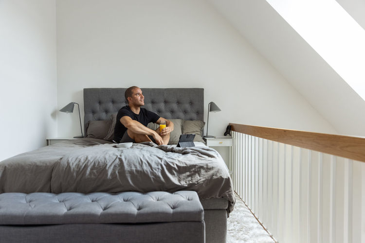 Side view of delighted male sitting on bed with glass of orange juice and browsing tablet while having breakfast in morning at home looking away