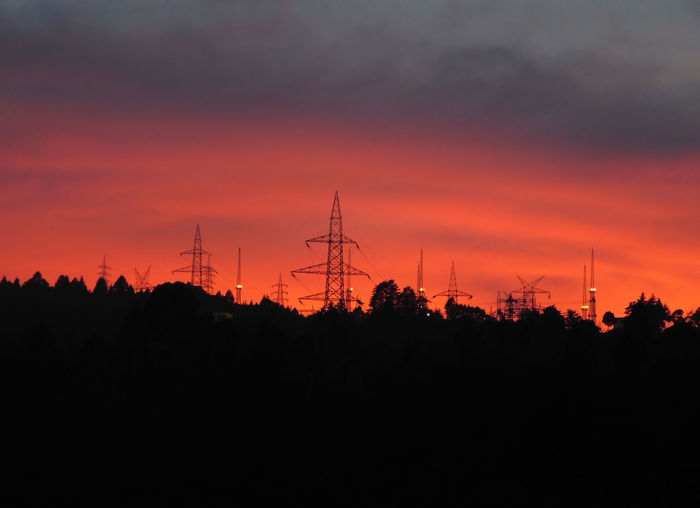 Silhouette electricity pylons by trees against romantic sky at sunset