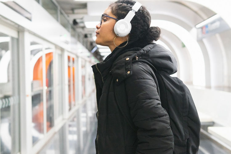 Side view of relaxed colombian female passenger in outwear and backpack listening to songs from playlist in wireless headphones while waiting for train on underground station