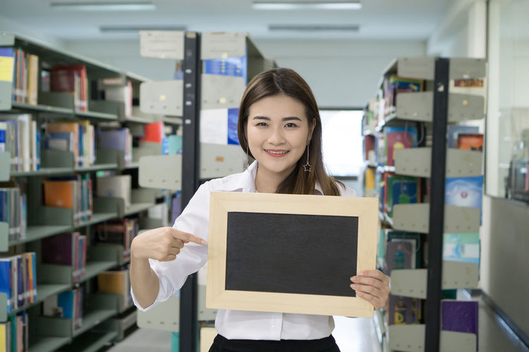 Portrait of smiling young woman pointing at writing slate in library