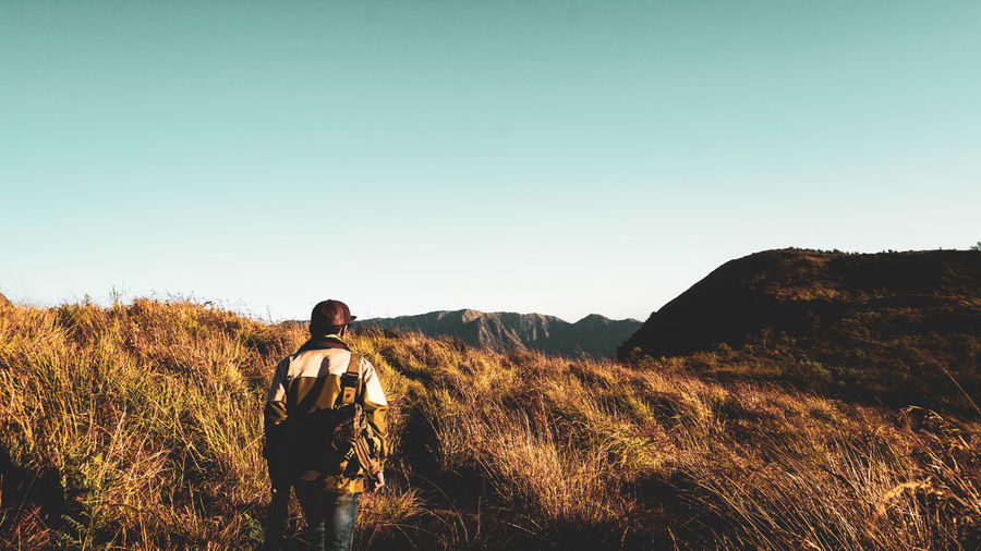 Rear view of man hiking on grassy mountain against clear sky during sunset