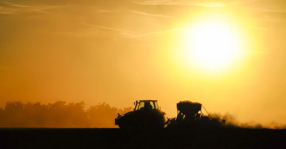 Silhouette of a tractor sowing seeds in a field in a cloud of dust against the background.