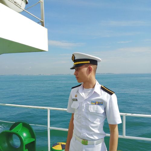Young navy officer standing on ship sailing in sea against sky