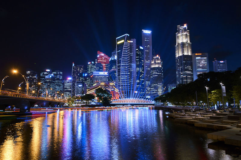 Illuminated bridge over river by buildings against sky at night