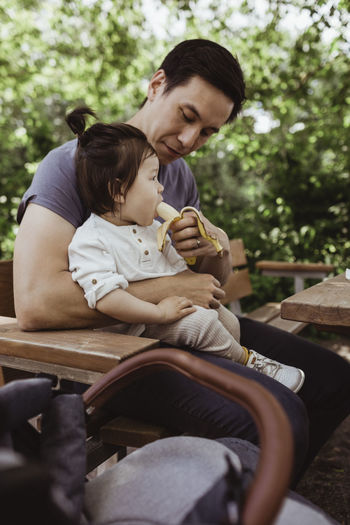 Father feeding banana to male toddler while sitting in park