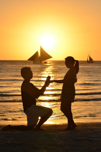 Man proposing woman at beach against orange sky during sunset