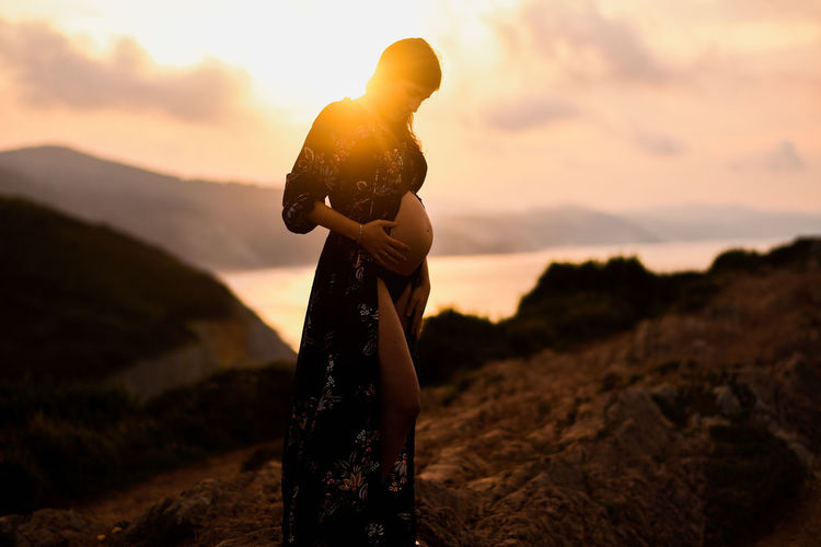 Side view of pregnant female standing alone on hill and dreaming about future at sunset time