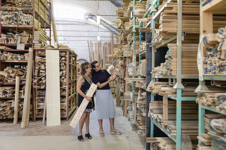 Women choosing ornate wood panel while standing in warehouse at factory
