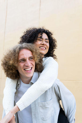 Cheerful man giving piggyback ride to diverse female friend both with curly hair laughing loud
