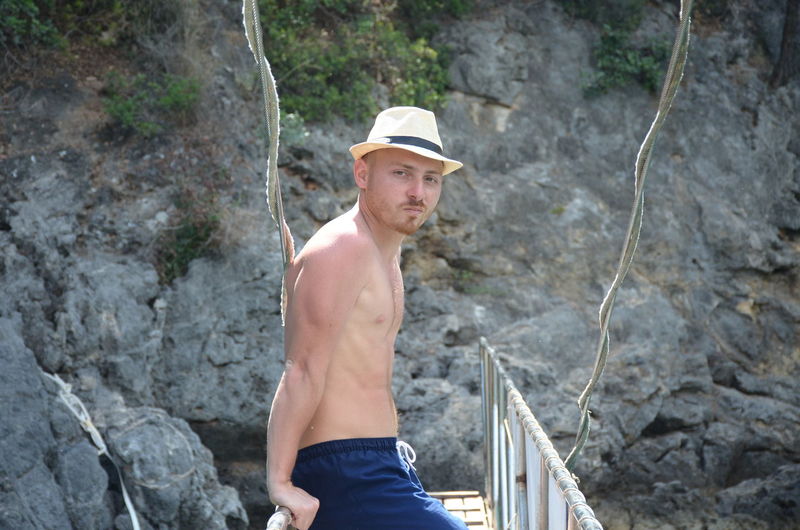 Portrait of shirtless man standing on bridge against rock formation