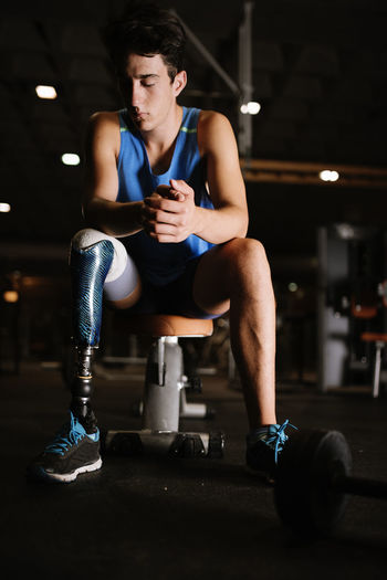 Full length of young man with prosthetic leg sitting on bench in gym