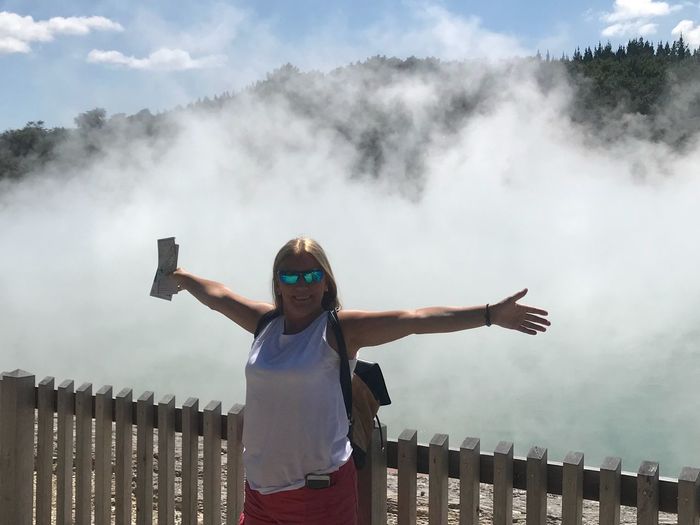 Portrait of mature woman with arms outstretched standing by fence against smoke erupting hot spring