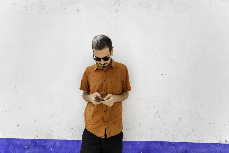 Young man wearing sunglasses using smart phone standing against wall