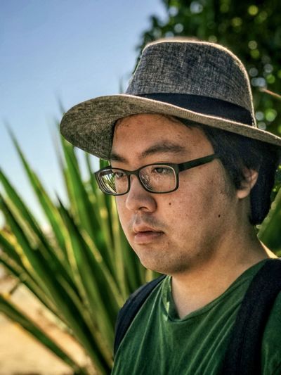 Close-up portrait of young man wearing eyeglasses and fedora against spiky green plant and blue sky.
