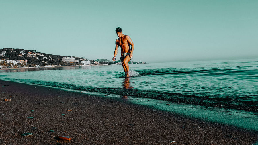 Full length of shirtless man standing at beach against sky